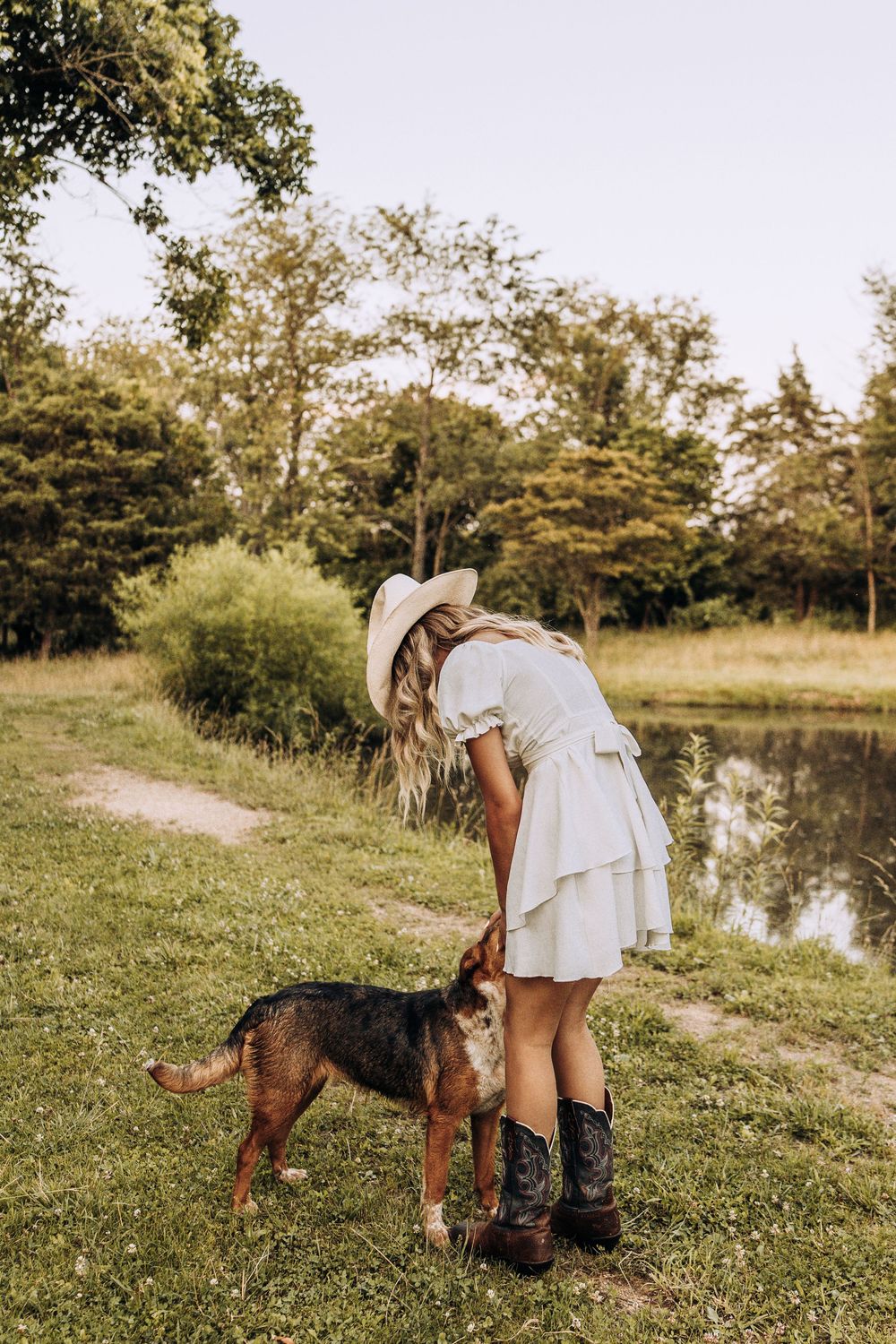 Blonde woman in white dress and cowboy boots bending to pet a dog near a peaceful pond.