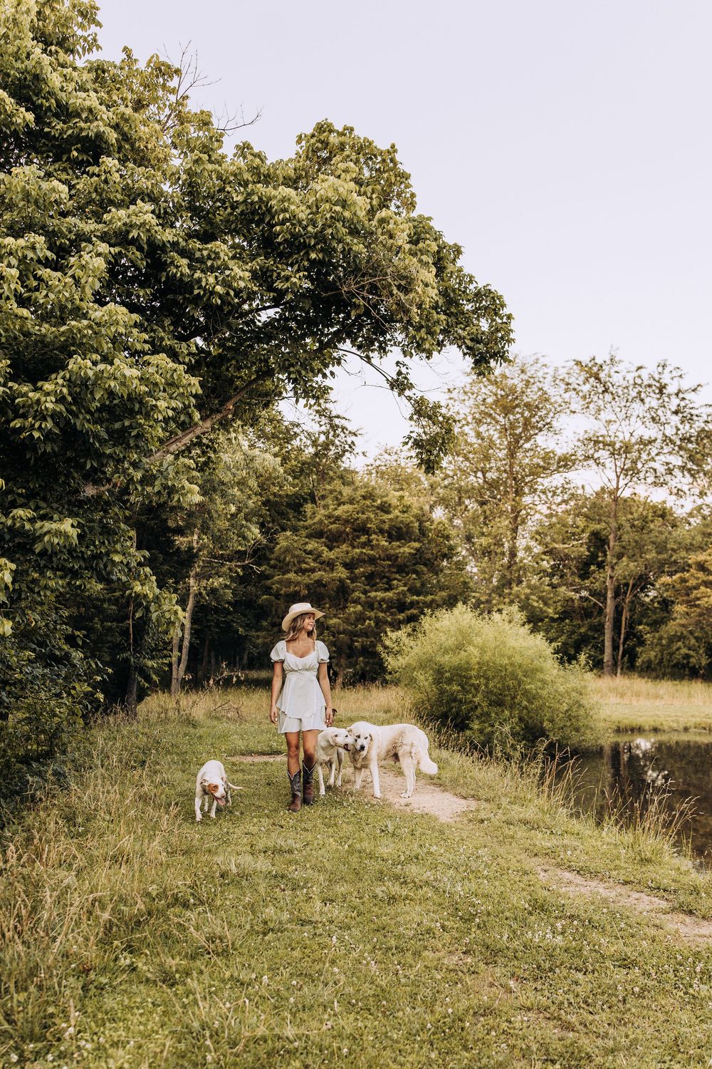 Woman in white dress and cowboy hat walking three dogs along a lush green path by a pond.