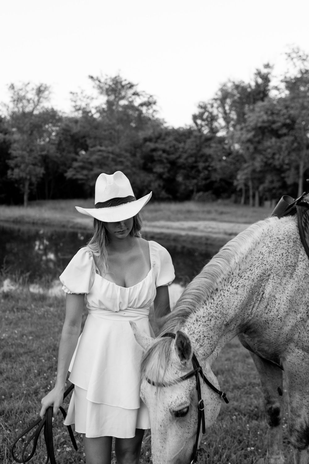 Black and white photo of woman in white dress and cowboy hat standing beside a grazing horse.