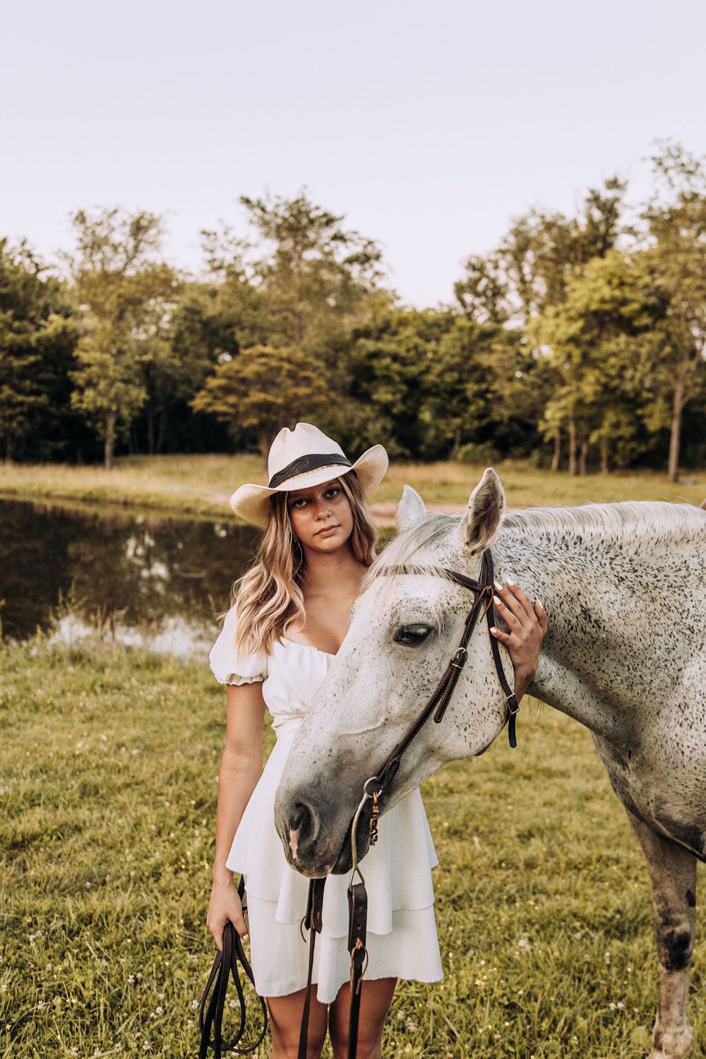 Woman in white dress and Panama hat stands beside a gray horse in a green field.