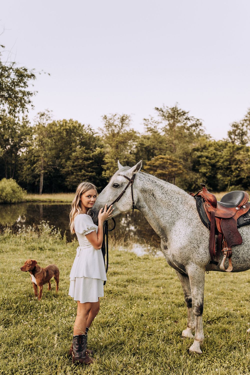 Young woman in white dress and cowboy boots posing with a gray saddled horse by a pond.