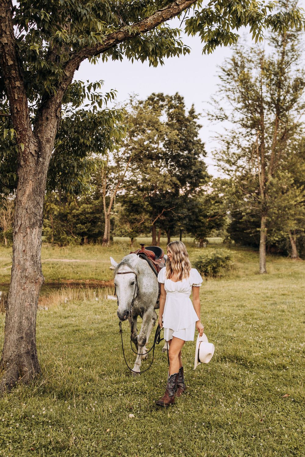 Woman in white dress and cowboy boots walks with a gray horse on a lush green farm.