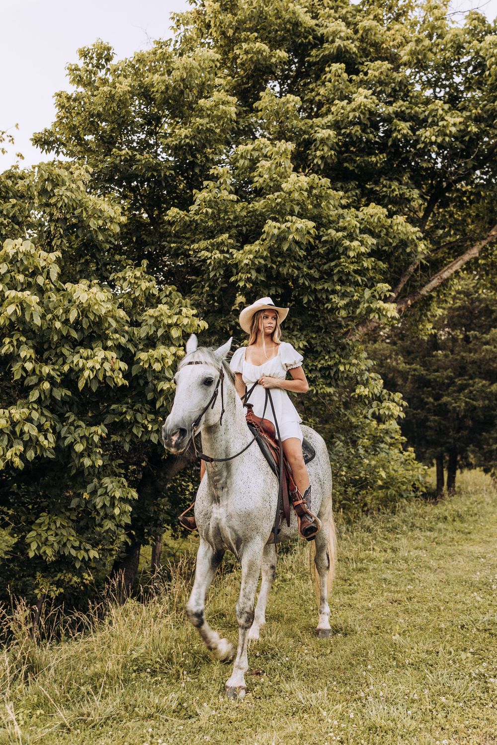 Woman in white dress and cowboy hat rides a gray horse through a lush green trail.