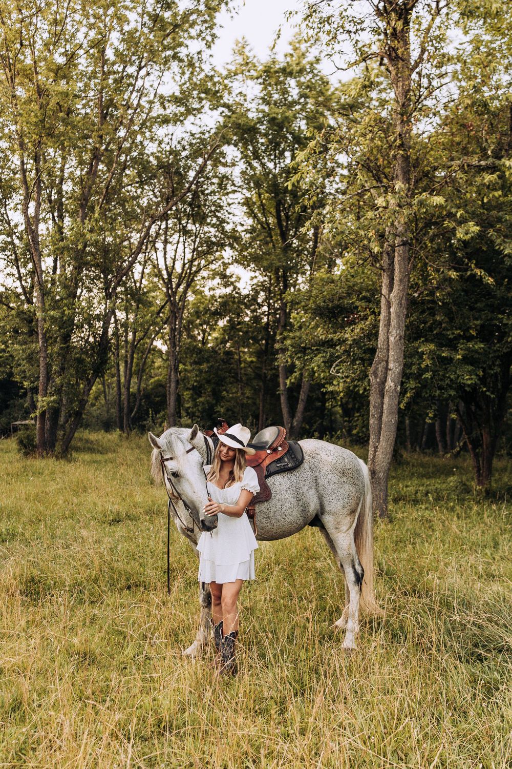 Woman in white dress and cowboy hat stands beside a saddled gray horse in a forest meadow.