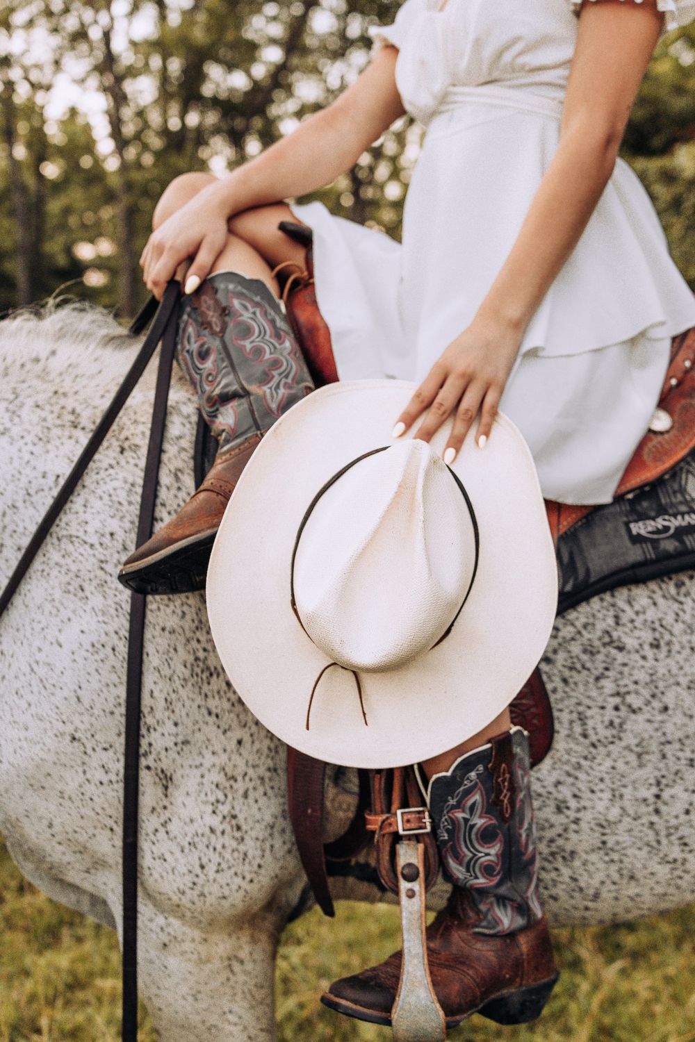 Close-up of cowboy boots, white hat, and reins on a gray horse with a woman in white dress.