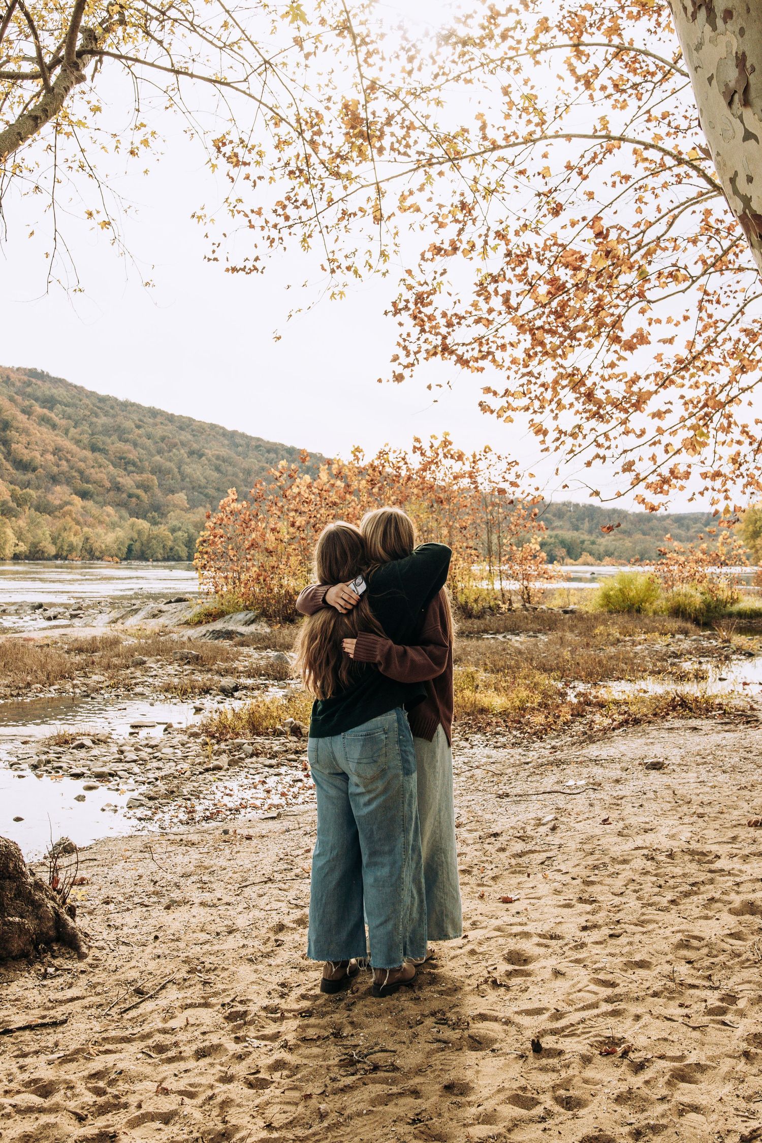 Two women embrace while standing on a sandy riverbank surrounded by autumn foliage and rolling hills.