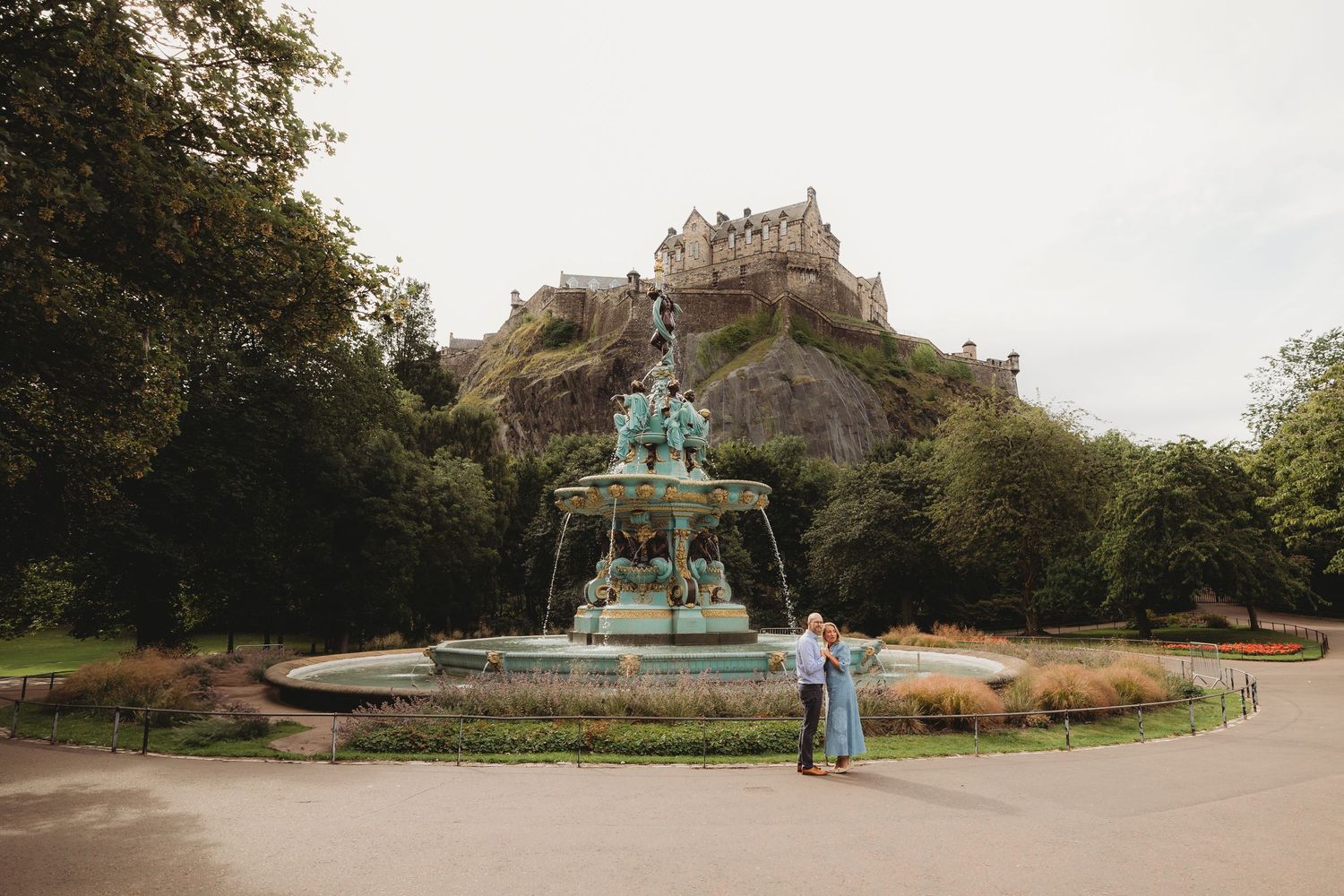 A couple shares a romantic moment in front of Edinburgh Castle and Ross Fountain in Princes Street Gardens.