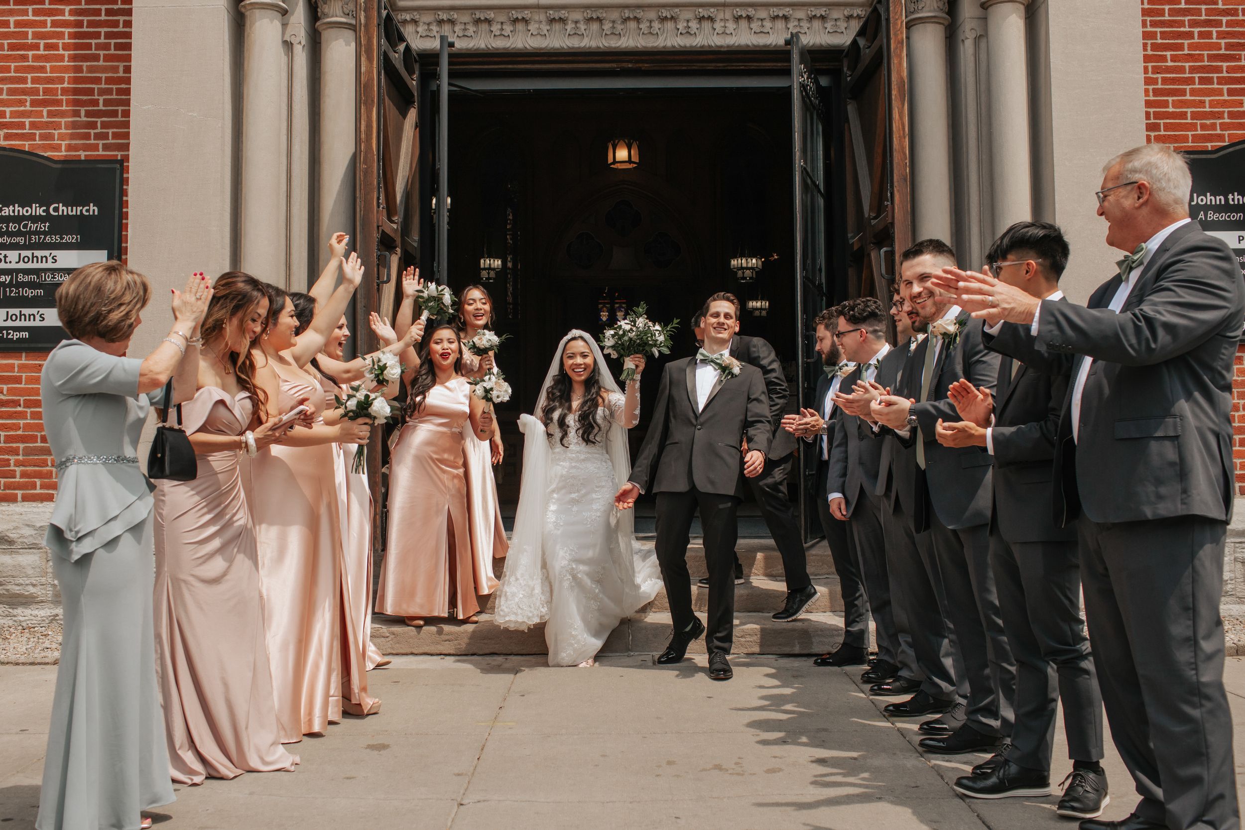Wedding party members in blush pink dresses and navy suits form an aisle outside a brick church entrance.