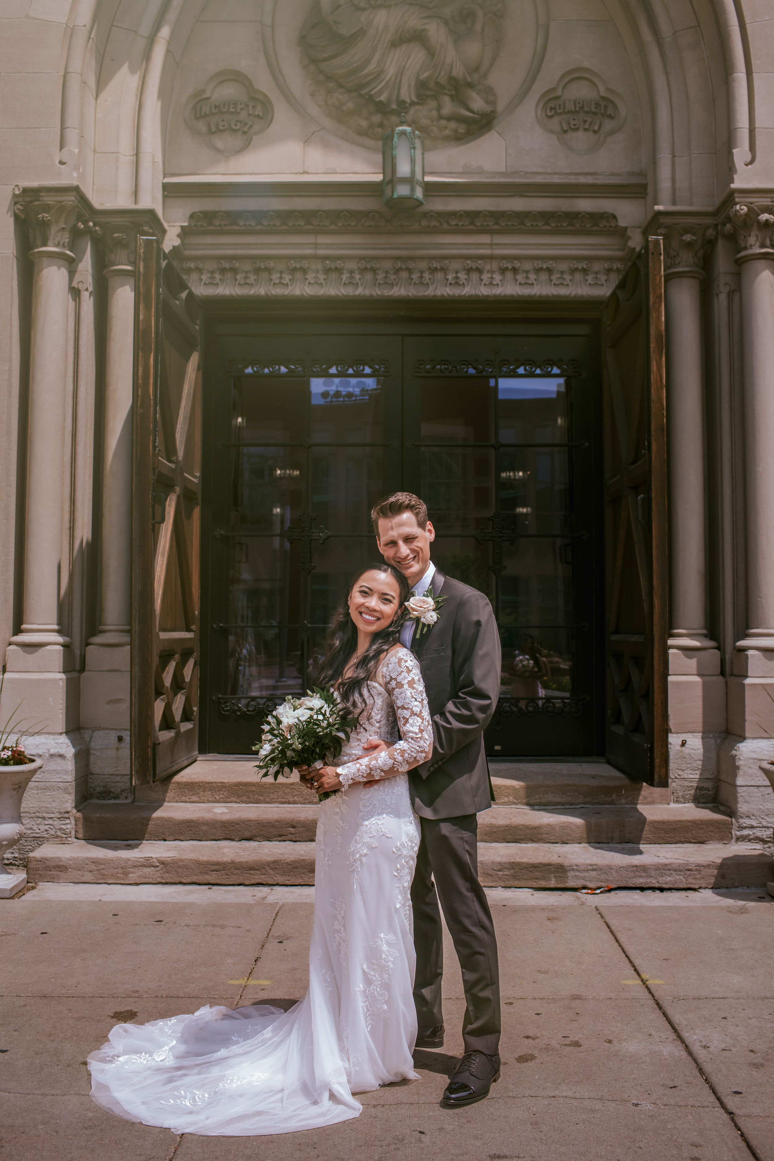 A couple in wedding attire shares a romantic moment outside a historic building with ornate architecture.