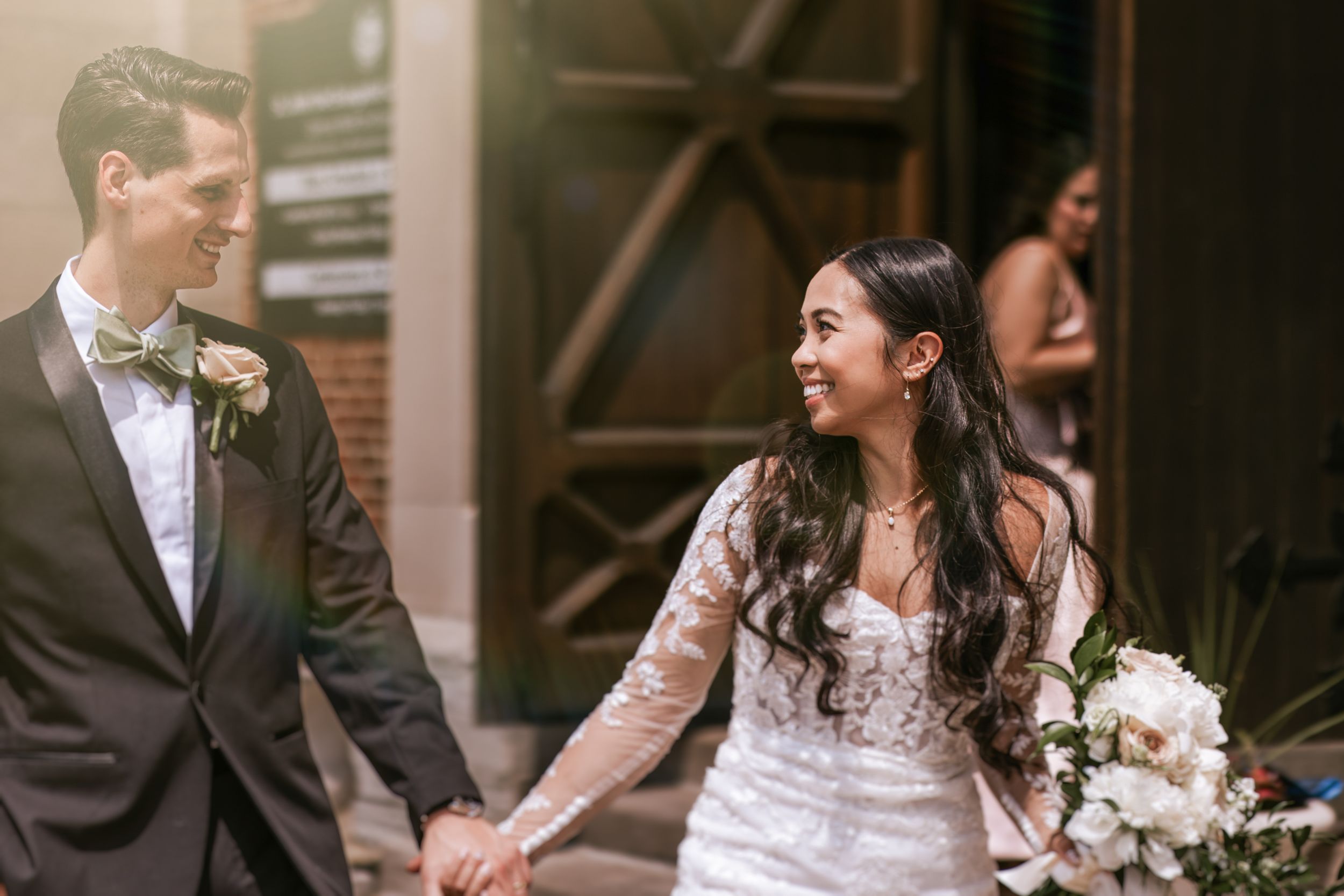 A joyful bride and groom hold hands and share a candid moment outside a rustic brick building.