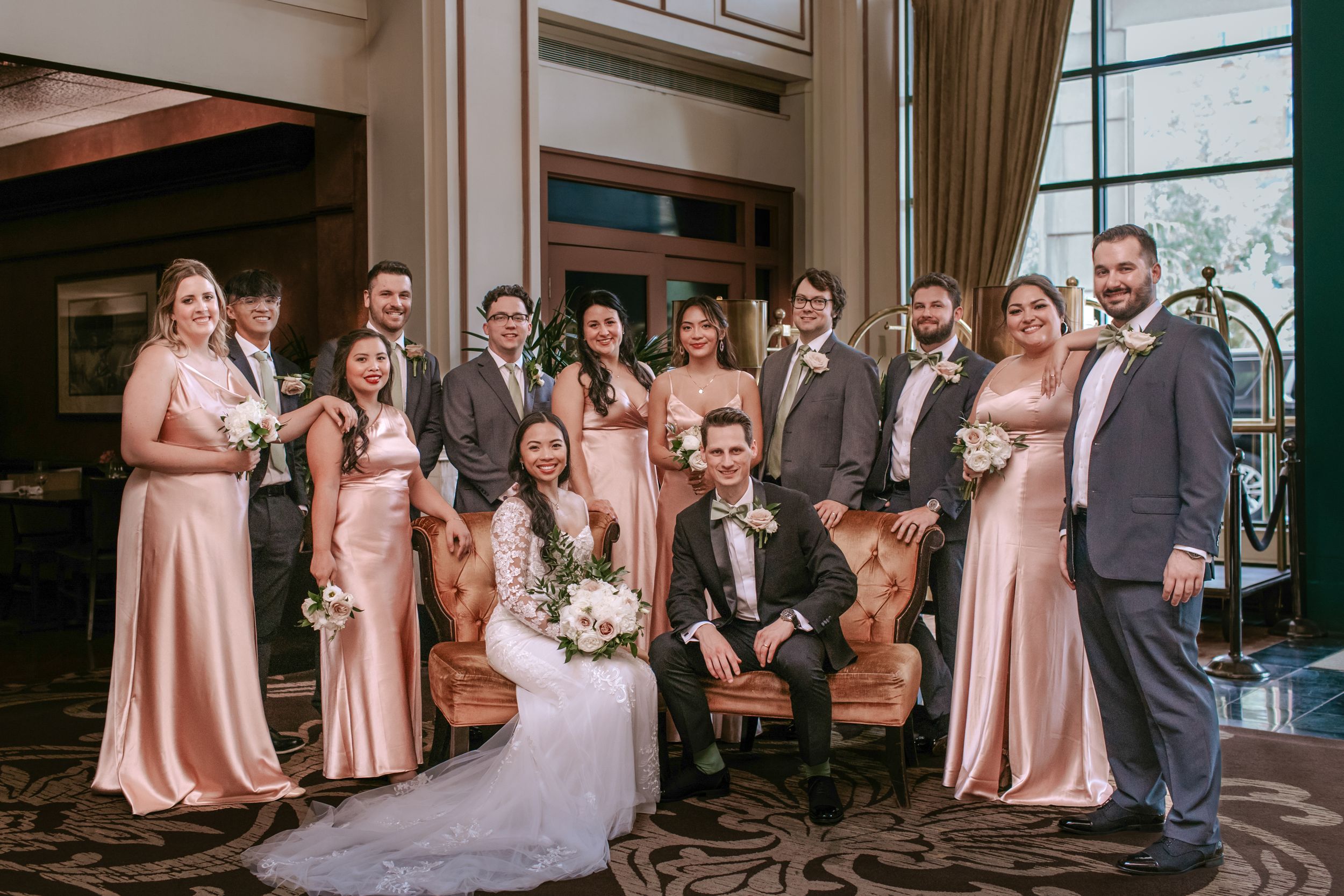 A wedding party poses together in formal attire with bridesmaids wearing blush pink dresses and groomsmen in dark suits.