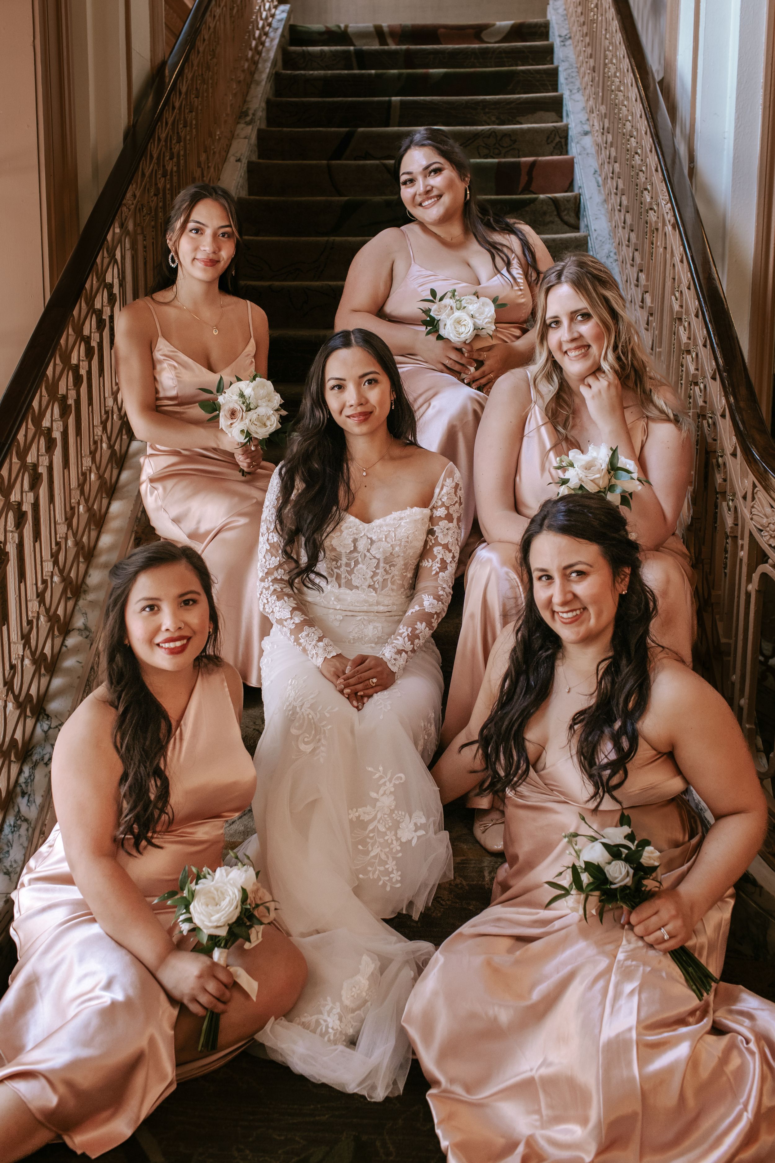 Wedding party in blush pink dresses pose together on elegant wooden staircase with white bouquets.