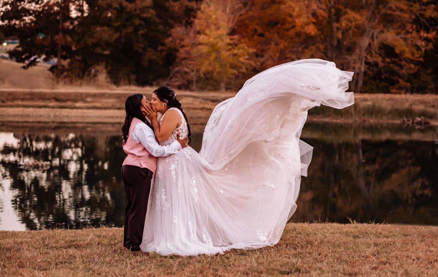 A romantic couple embraces in multiple poses near a lake during autumn, with the bride's white dress flowing in the breeze.