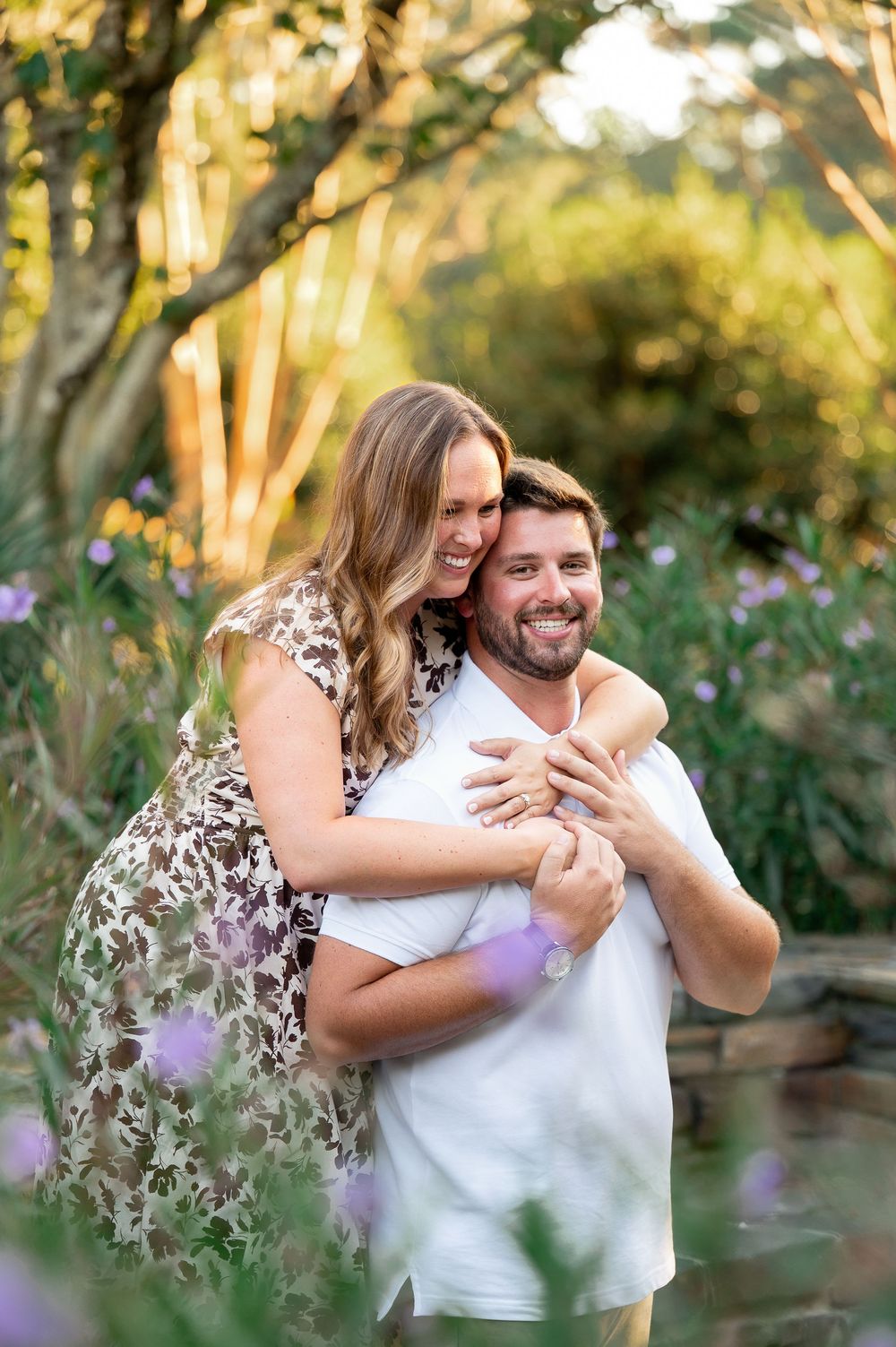Couple embraces and laughs together in garden setting with soft purple flowers.