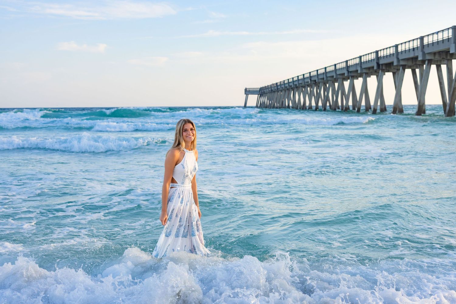 A person in a white dress walks through the turquoise ocean waves near a wooden pier at sunset.