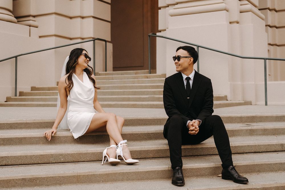 Bride and groom sit on elegant stone steps in formal wedding attire during outdoor photo session.