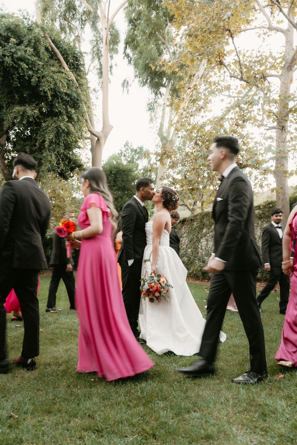Wedding party in formal attire stands in line against backdrop of autumn trees during outdoor ceremony.