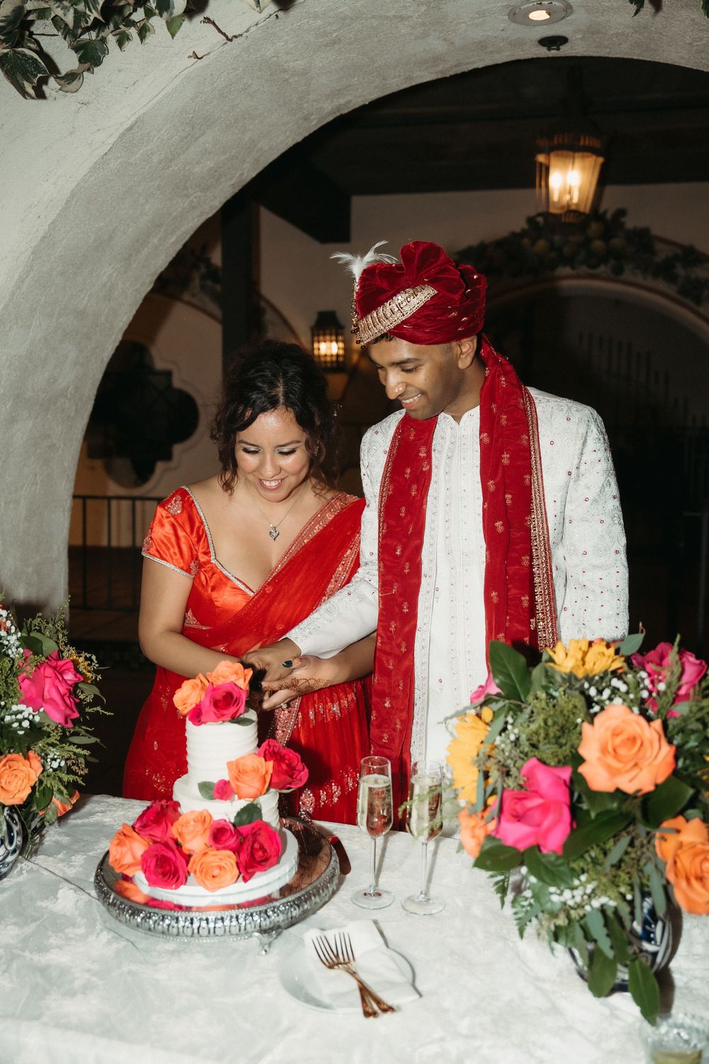 A couple in red and white traditional wedding attire share a slice of wedding cake at their reception table.