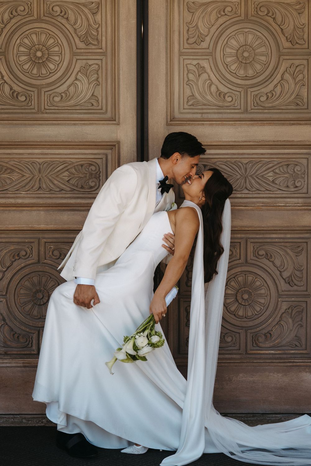 A romantic wedding couple in white attire share a kiss against an ornate vintage door with decorative patterns.