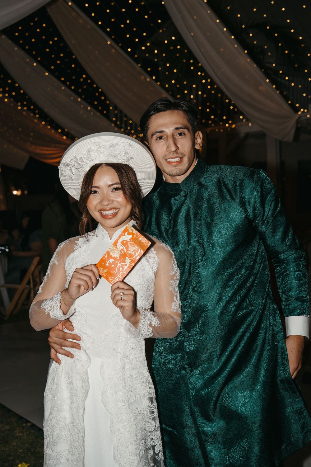 A couple in wedding attire holds up orange envelopes during a reception under twinkling lights.