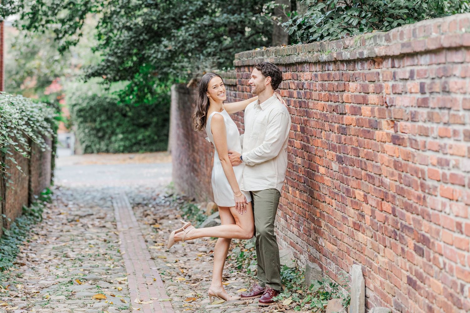 A romantic couple strolls along a brick wall pathway surrounded by lush greenery in an outdoor garden setting.