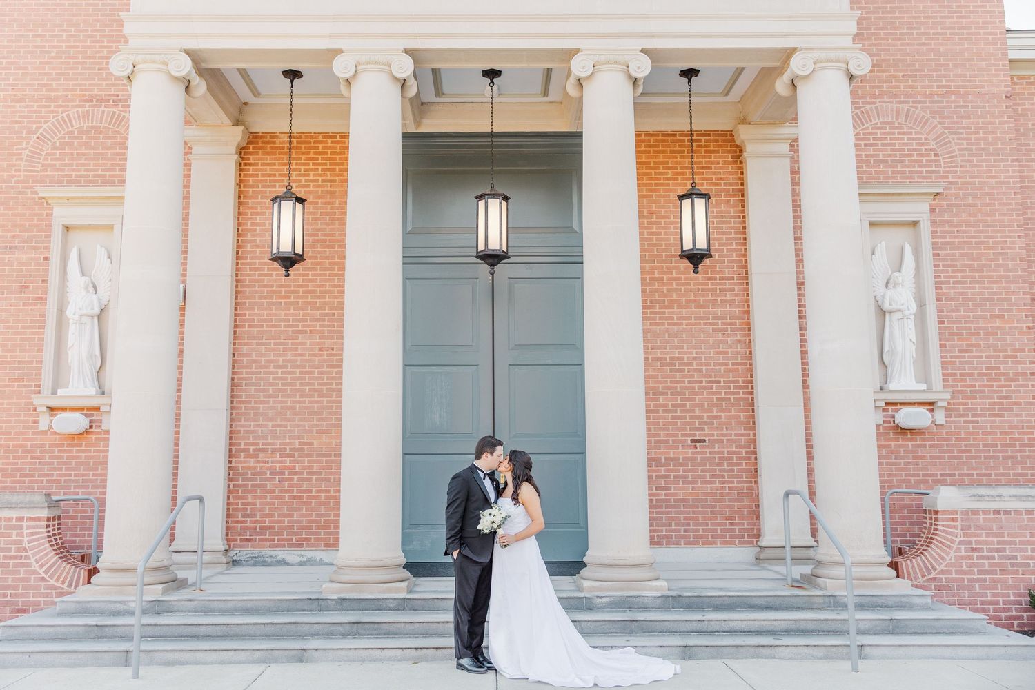A bride and groom share a romantic moment in front of an elegant brick building with white columns and lanterns.