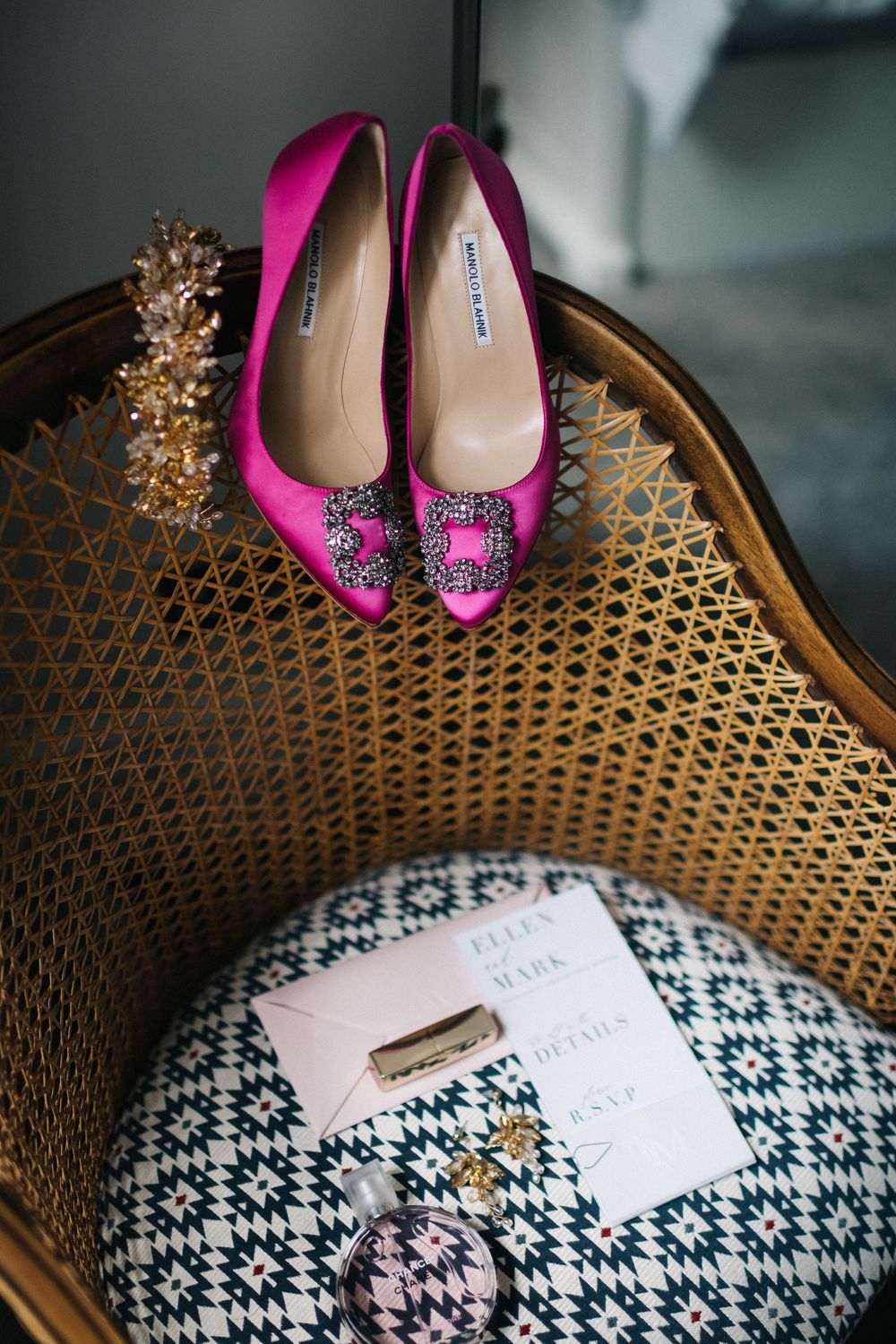 Pink bridal shoes and wedding invitation displayed on a vintage wicker chair with decorative pattern.