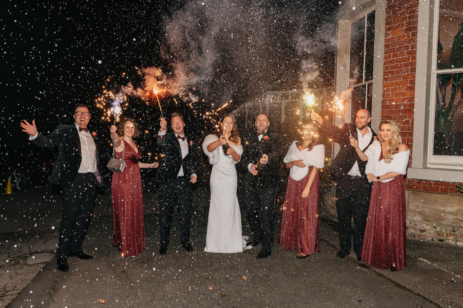 A wedding party celebrates with sparklers outside a brick building at night.