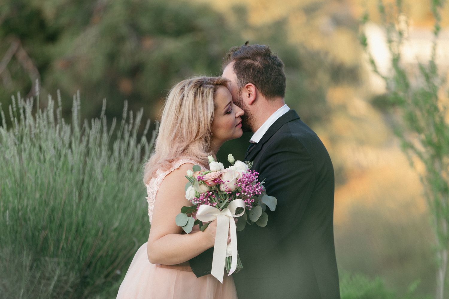 A romantic wedding couple shares an intimate kiss surrounded by tall grass during a golden sunset photo session.