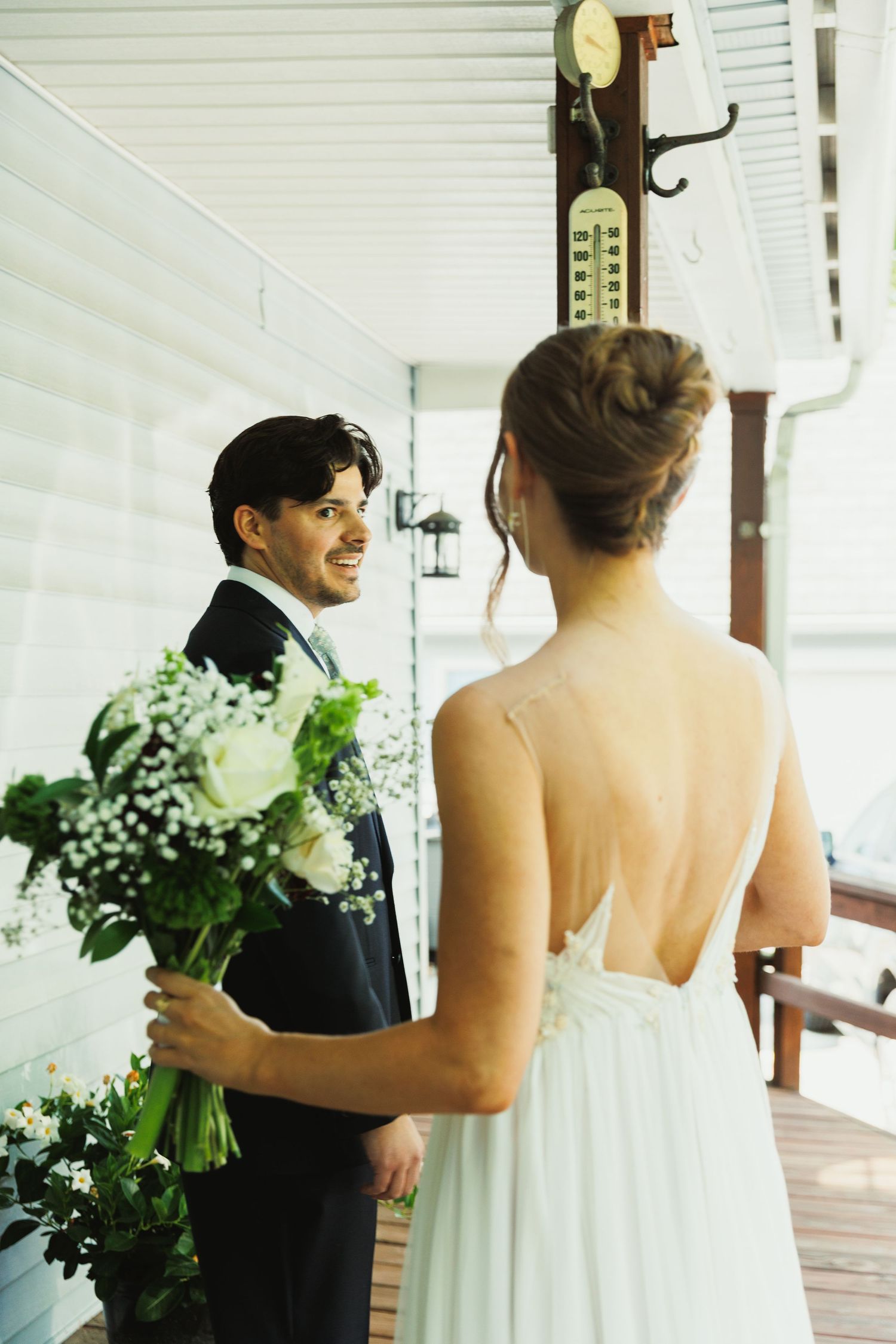 A couple shares their first look moment on a white porch holding floral bouquets.