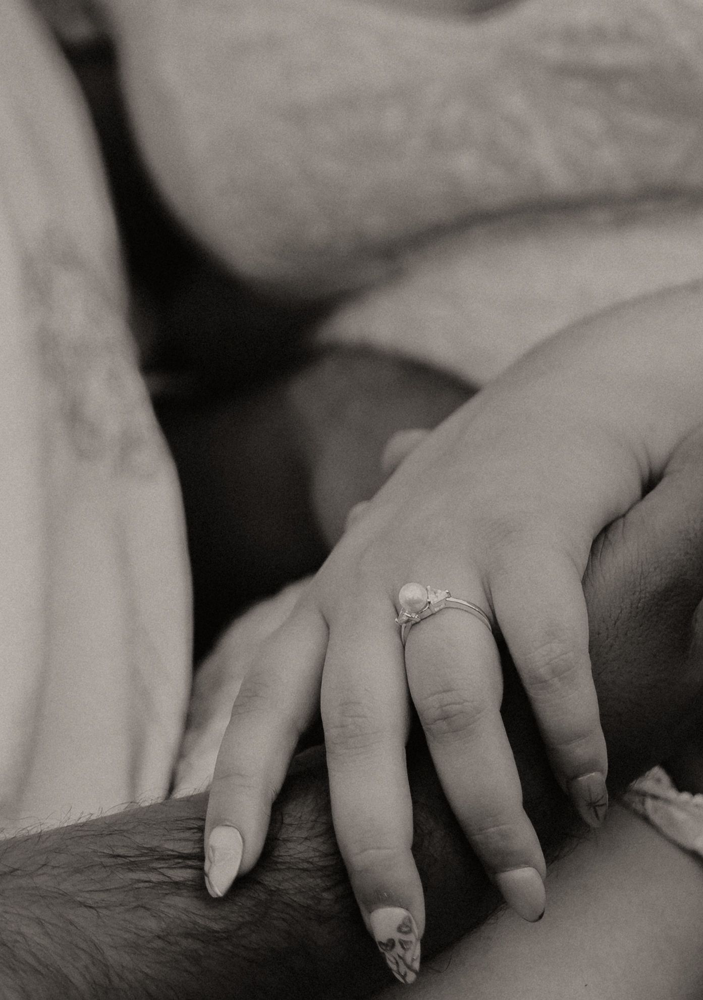 Black and white close-up photo of hands with an engagement ring resting on soft fabric.