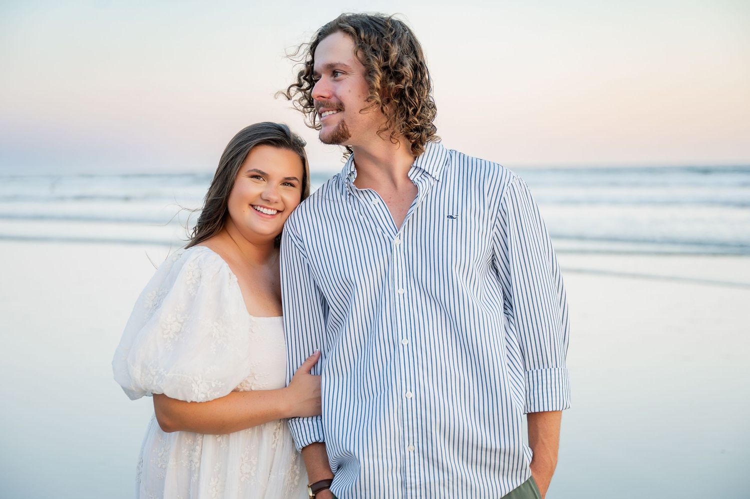 A couple embraces on a sandy beach with ocean waves in the background.