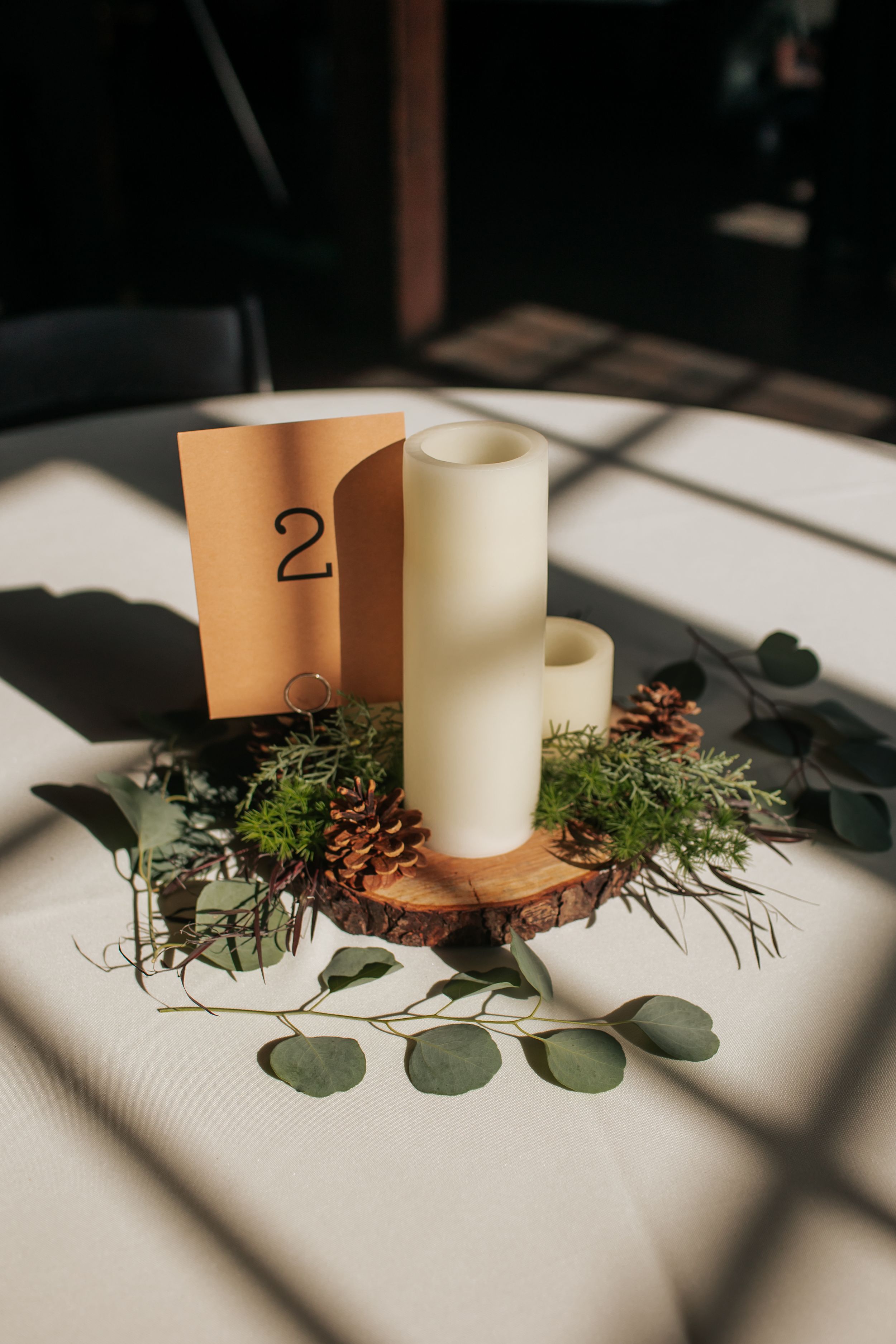 White candles arranged with eucalyptus leaves and pine cones create an elegant table centerpiece in natural light.