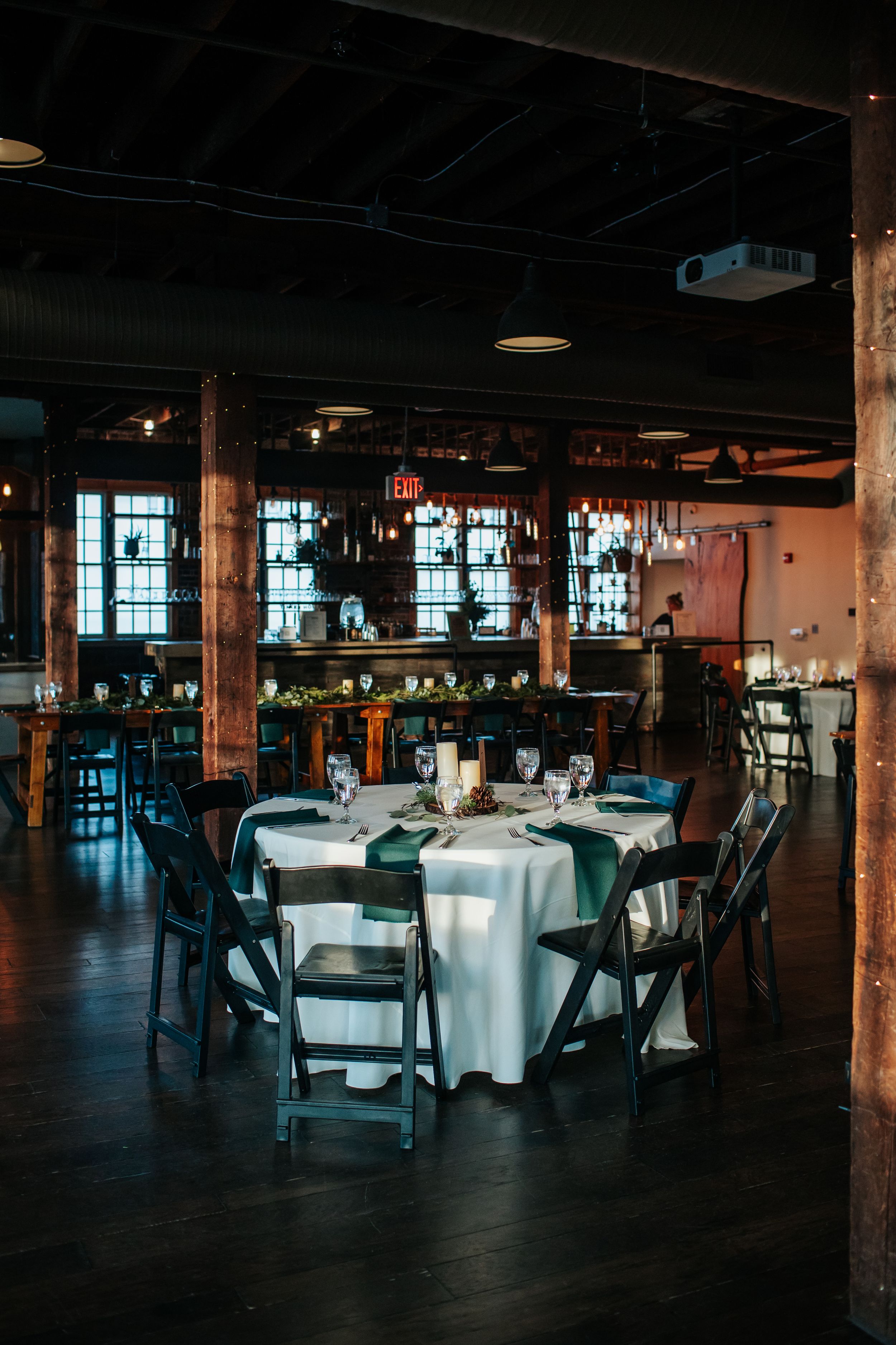 Elegant event space with round tables, white linens, and industrial-chic decor featuring exposed beams and mood lighting.