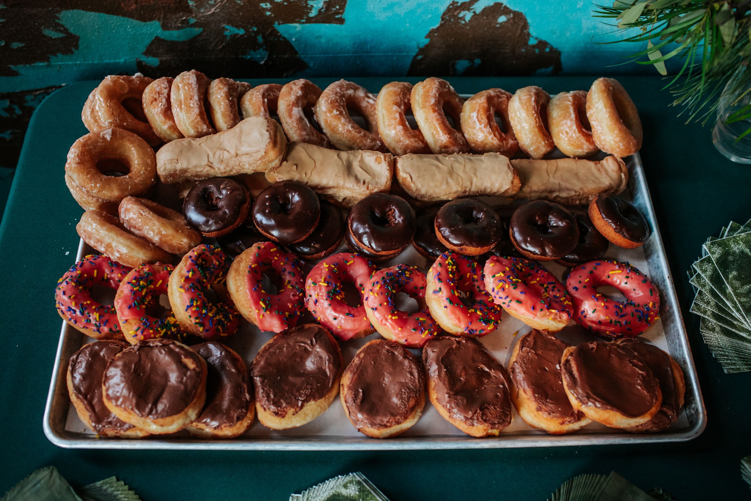 Rows of colorful glazed and frosted donuts arranged neatly on a metal tray against a teal background.
