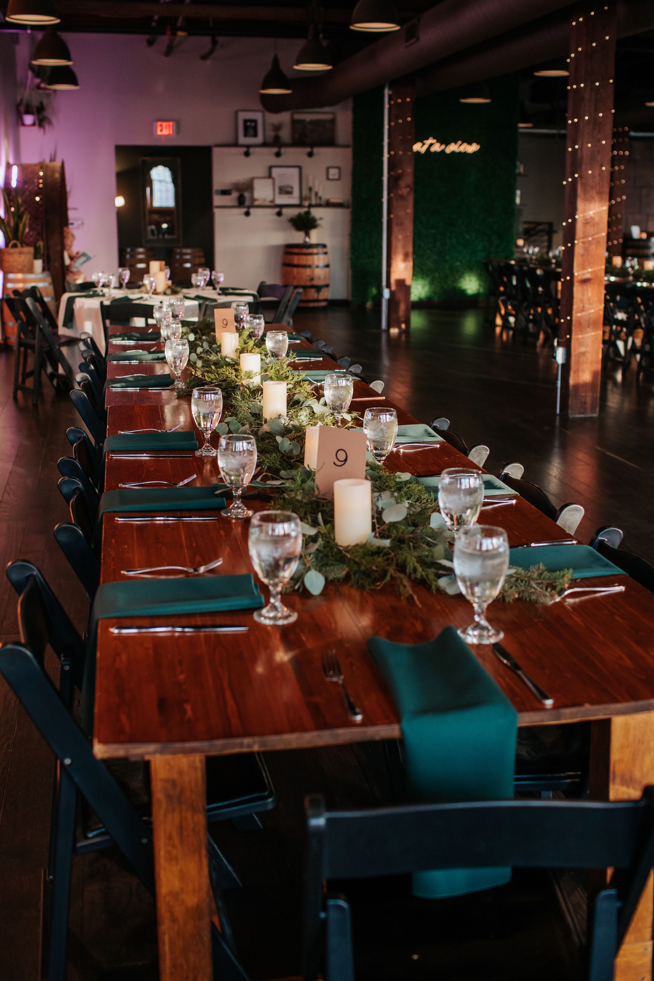 Elegant table setting at a restaurant with wooden tables, teal chairs, and greenery centerpiece decorations.