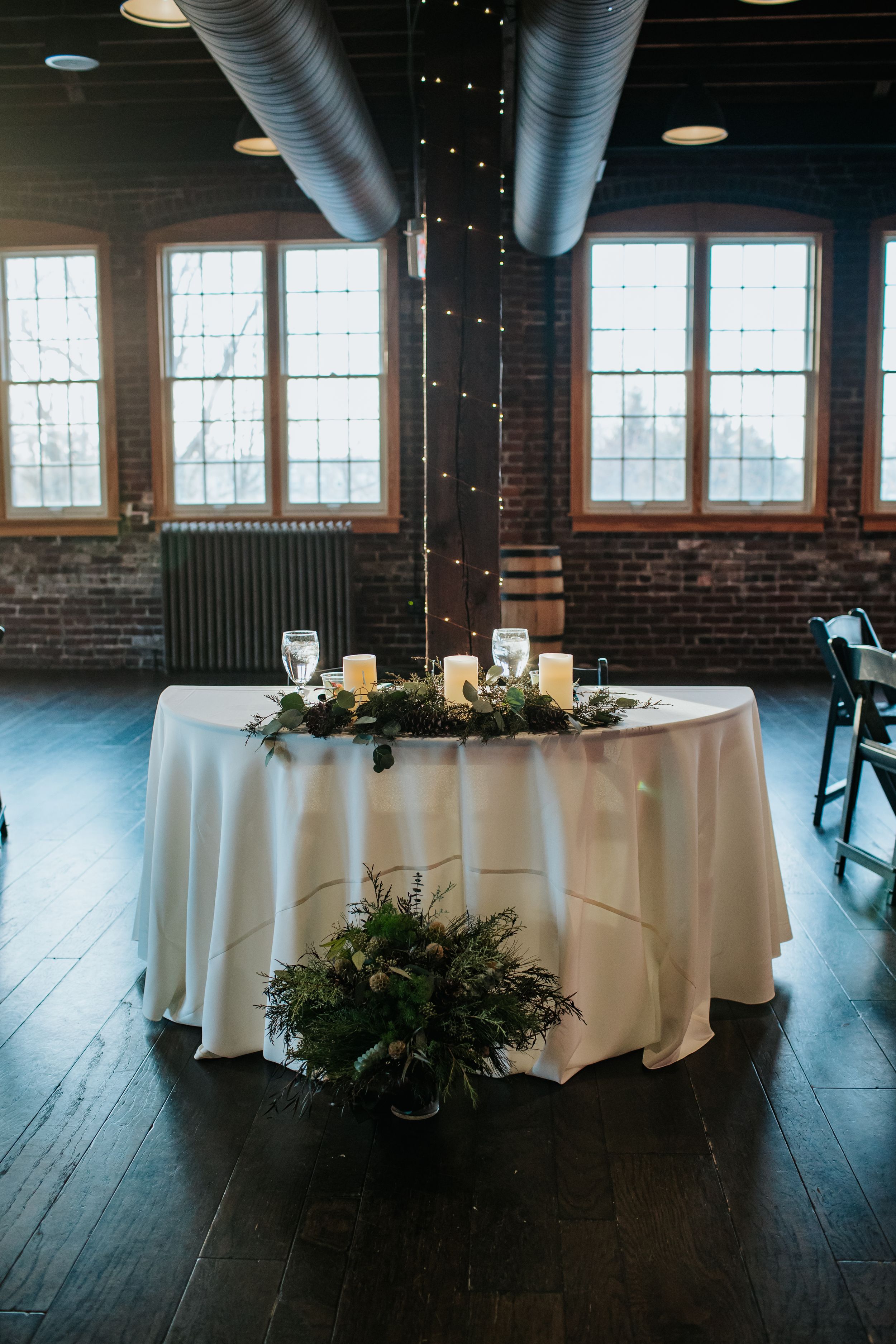 Round banquet tables with white tablecloths and greenery centerpieces set up in an industrial-style venue with exposed brick walls.