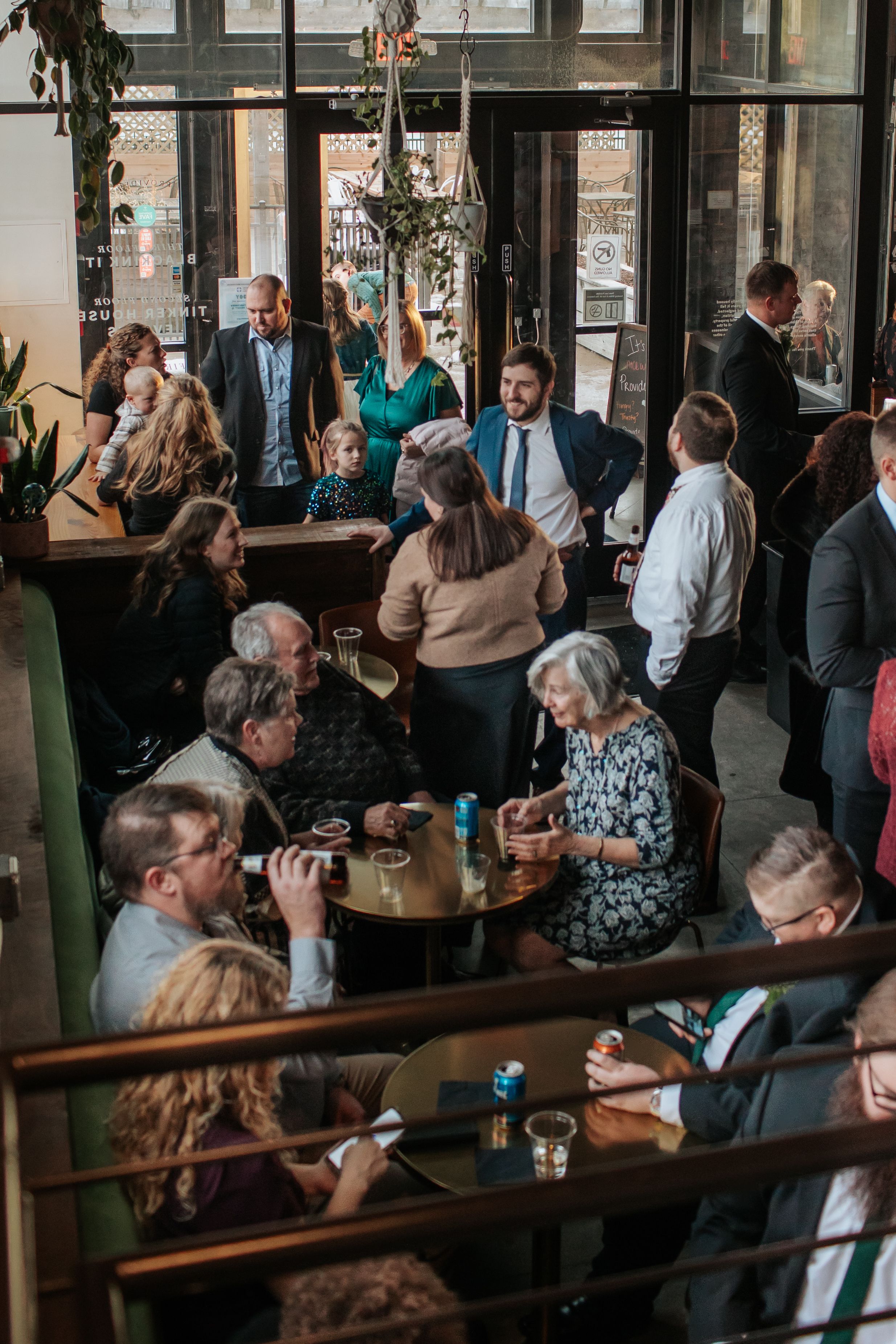 A crowded restaurant scene with people gathered at tables and standing, featuring large windows and modern decor.