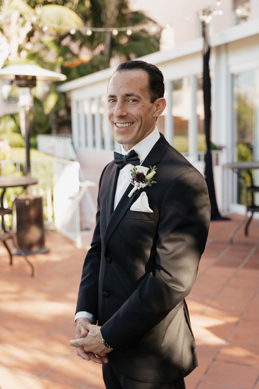 Groom in black tuxedo smiling while reading a letter during outdoor wedding ceremony.