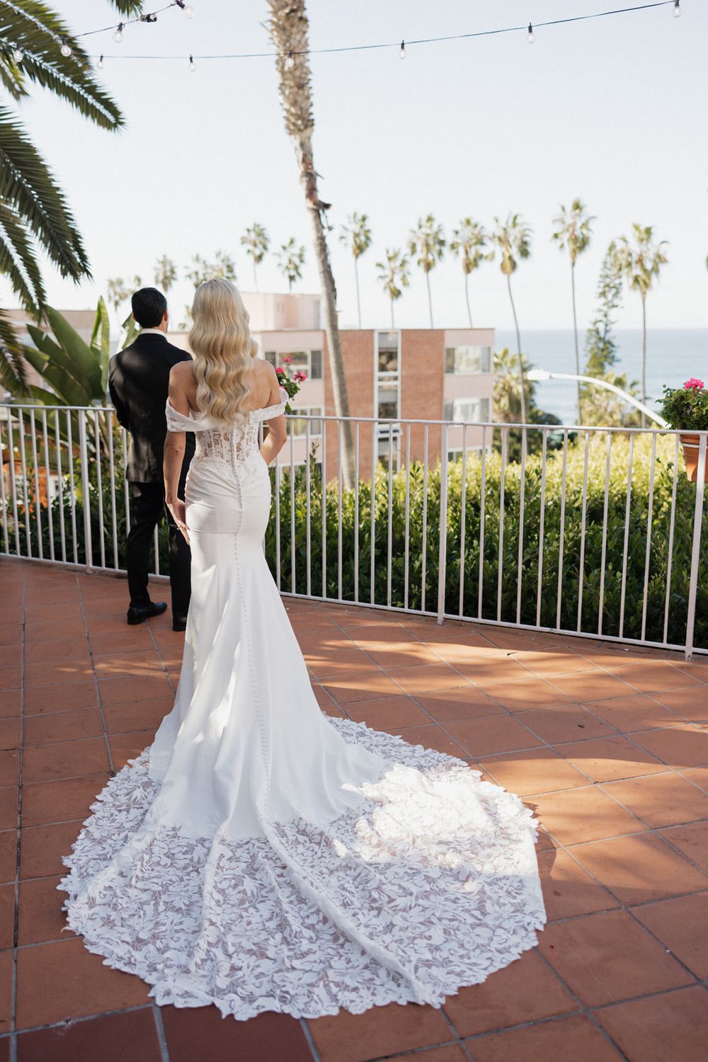 A bride in a white lace wedding dress with a dramatic train stands on a terrace overlooking palm trees at sunset.