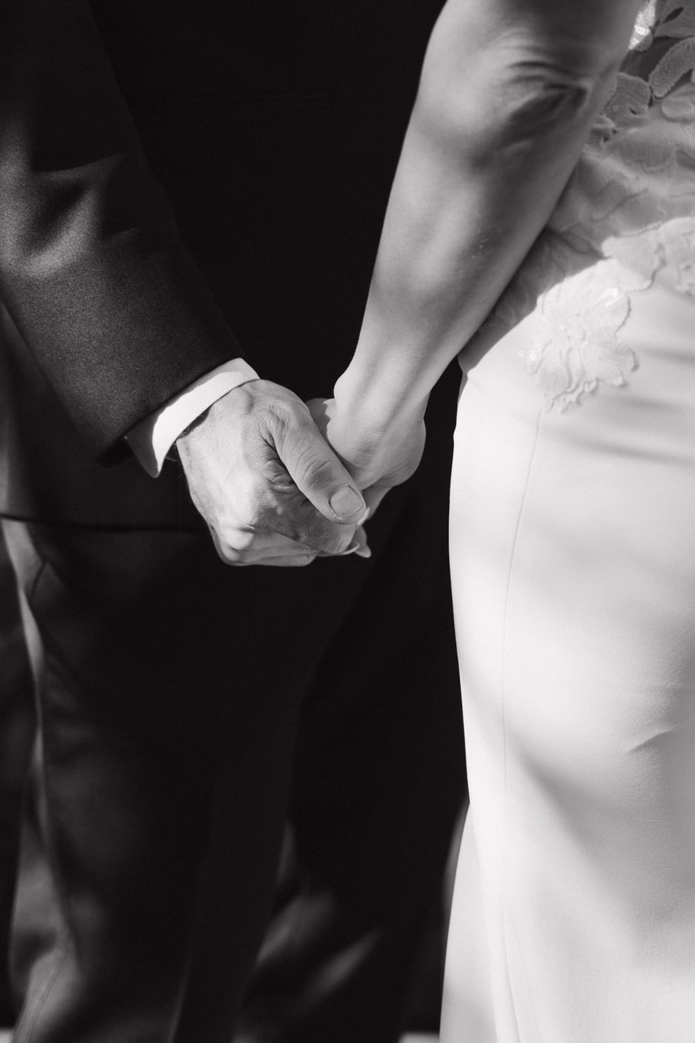 Black and white detail shot of newlyweds holding hands, showing the contrast between wedding dress and tuxedo.