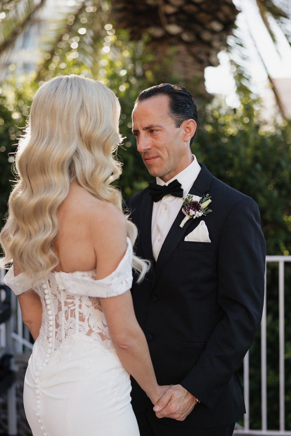 A bride in a white lace wedding dress and groom in black tuxedo share an intimate moment outdoors.