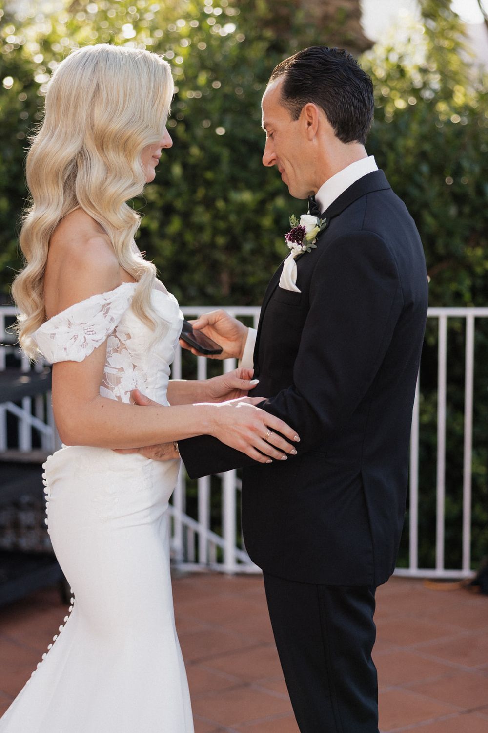 Couple exchanges rings during outdoor wedding ceremony with bride in off-shoulder gown and groom in black tuxedo.