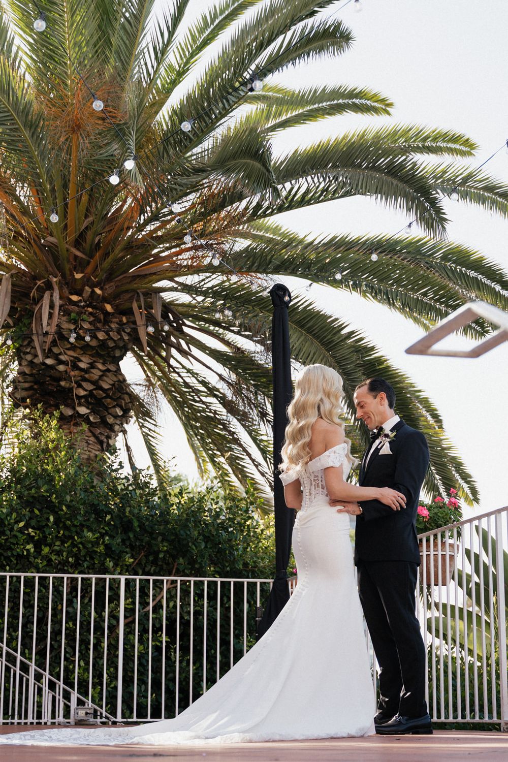 A couple shares an intimate moment on a terrace with a palm tree and white railing during their wedding celebration.