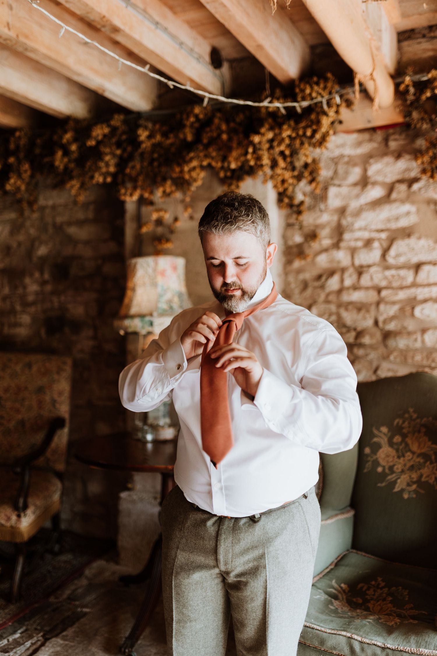 Groom in a white shirt and gray vest stands in a rustic stone-walled room with exposed wooden beams at Lyde Court, Hereford.