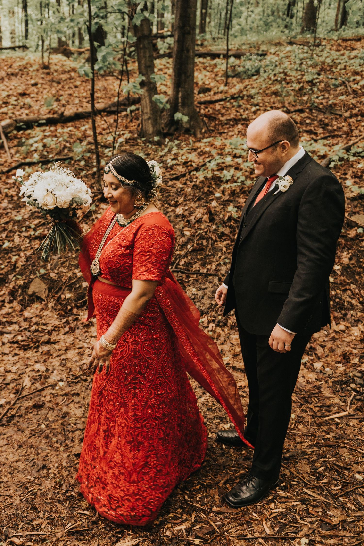 A couple in formal attire walks through an autumn forest with fallen leaves, the bride wearing a stunning red dress.