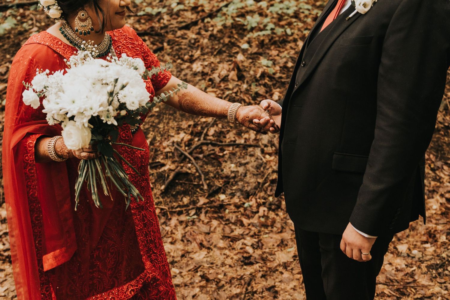 Wedding couple in formal attire poses with white bouquet among fallen autumn leaves in forest setting.