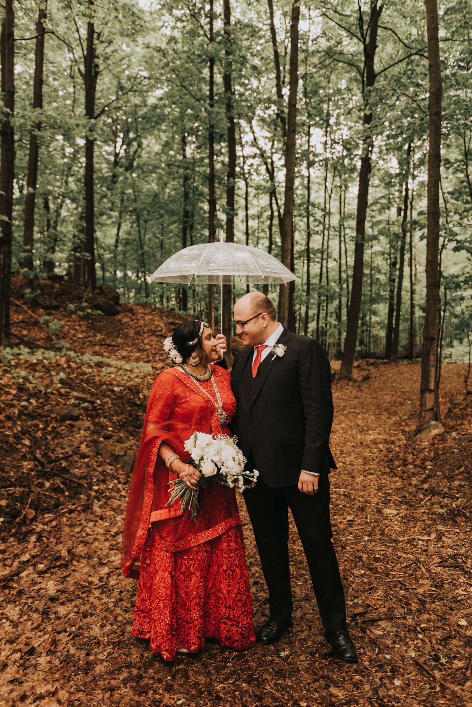 A couple shares a romantic moment under a clear umbrella in an autumn forest with fallen leaves.