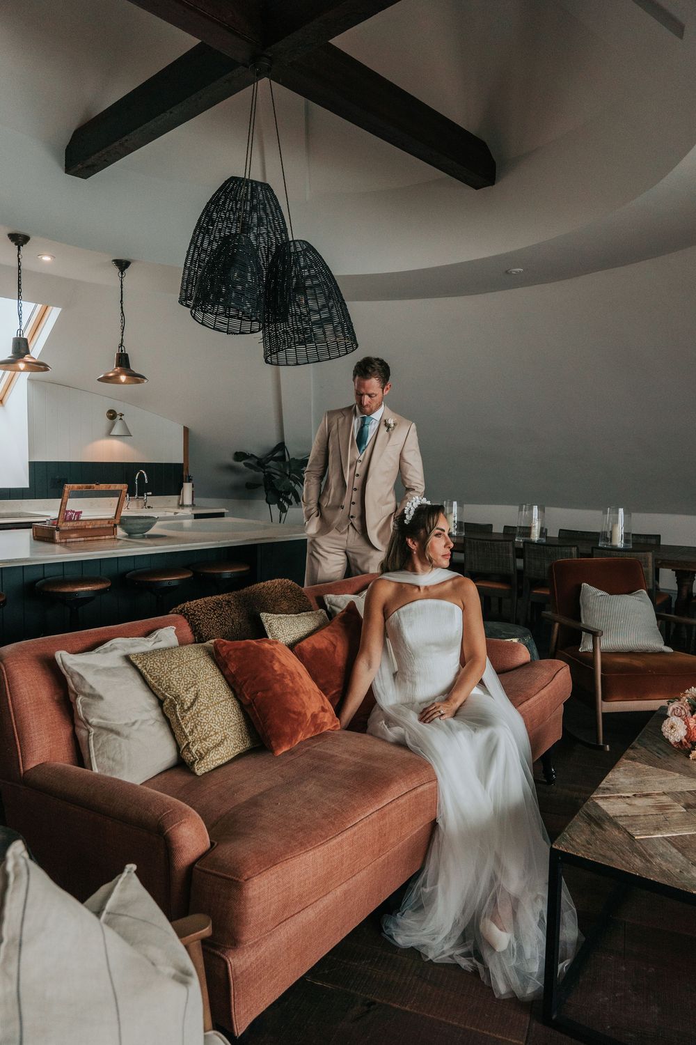 Editorial-style portrait of a bride seated on a sofa with her groom standing behind her in a Gara Rock Hotel suite, photographed during a re