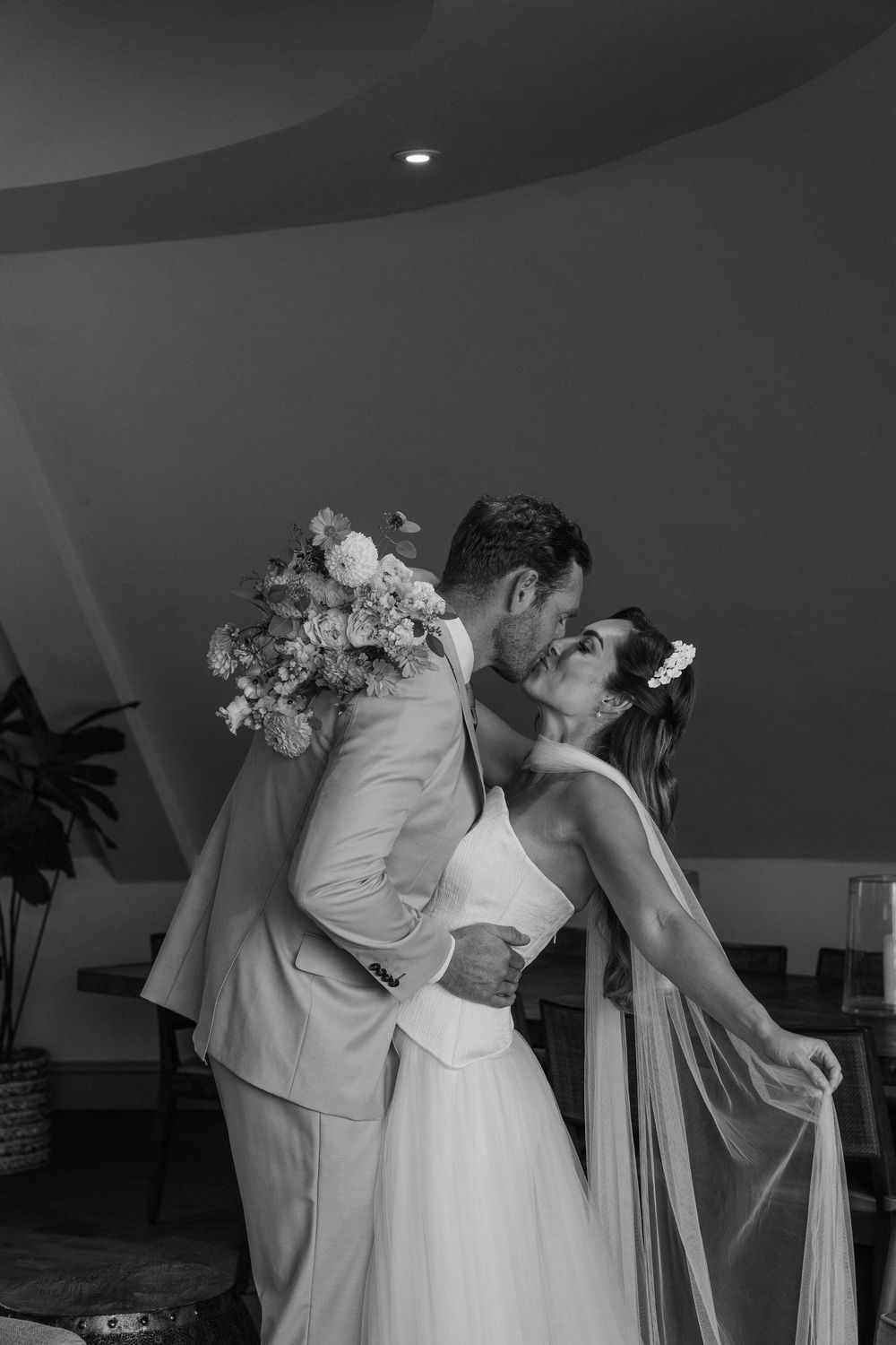 Black and white editorial wedding portrait of a bride and groom kissing indoors at Gara Rock Hotel, with soft movement and florals captured